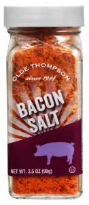 A clear jar of Olde Thompson bacon salt. The label features a pig silhouette on a purple background. The text reads Olde Thompson since 1944 Bacon Salt. The jar contains 3.5 oz (99g) of product.