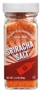 A transparent glass jar of Olde Thompson Sriracha Salt with vibrant orange-red salt. The label reads Olde Thompson since 1944, with a hot sauce bottle icon and Net Wt. 3.3 oz (94g) written below.