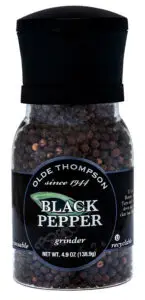 A clear plastic bottle of Olde Thompson black pepper with an attached grinder. The bottle contains whole peppercorns and has a black cap. The label includes text about the product and a recycling symbol.