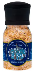 A clear bottle of Olde Thompson California Garlic & Sea Salt with a black grinder top. The blue label features yellow and white text, indicating its a 8.8 oz (249.4g) recyclable product.