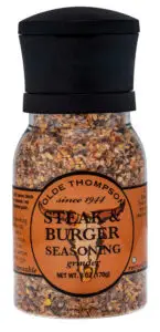 A clear plastic grinder bottle of Olde Thompson Steak & Burger Seasoning, featuring a black cap and an orange label. The label includes seasoning details and the net weight of 6 oz (170g).