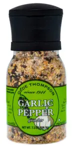 A clear plastic grinder bottle with a black top contains a blend of garlic and pepper. The green label reads Olde Thompson Garlic Pepper Grinder and displays additional text about the product. Bottle net weight is 7.3 oz (206.9g).