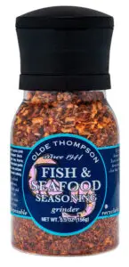 A bottle of Olde Thompson Fish & Seafood Seasoning with a grinder top. The label is dark blue with white and pink text, detailing that it contains 5.5 oz (156g) of seasoning. The bottle is clear, showing a mix of seasoning inside.