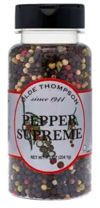 A clear plastic jar labeled Olde Thompson Pepper Supreme contains a mix of colorful peppercorns. The jar features a black lid and displays text indicating a net weight of 7.2 ounces (204.1 grams).