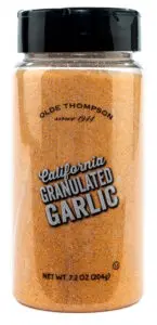 A clear plastic jar with a black lid, containing granulated garlic. The label reads Olde Thompson since 1944, California Granulated Garlic. Net weight is 7.2 oz (204g). The granules inside are golden brown.