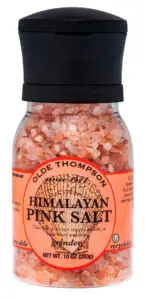 A clear grinder bottle filled with coarse pink salt crystals, labeled Olde Thompson Himalayan Pink Salt. The orange label includes text about it not supplying iodine and mentions that its recyclable. Net weight is 10 oz (283g).