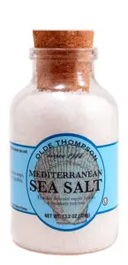 A clear jar with a cork lid containing coarse Mediterranean sea salt. The jar has a blue label with decorative text. It holds 13.2 oz (374g) of salt.