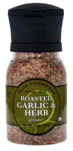 A clear grinder bottle labeled Olde Thompson Roasted Garlic & Herb, containing a coarse spice blend. The bottle has a black twist-top and green label, weighing 5.9 oz (167g), marked as recyclable and disposable.