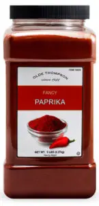 A large plastic container of Olde Thompson Fancy Paprika, featuring a label with an image of paprika powder in a bowl and a small red pepper. The container holds 5 lbs (2.27 kg) of paprika.