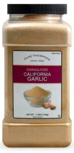 A large plastic container labeled Olde Thompson Granulated California Garlic with a net weight of 7 lbs (3.18 kg). The label features an image of a bowl of granulated garlic and a few garlic cloves.