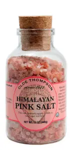 A clear glass jar with a cork stopper contains coarse Himalayan pink salt. The label reads Olde Thompson Himalayan Pink Salt and notes a weight of 12 oz (340g). The product is described for use in cooking and seasoning.