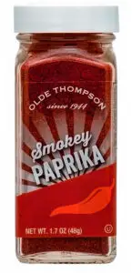 A clear glass jar of Olde Thompson smokey paprika. The label features a sunburst design behind the text, with a red chili pepper illustration at the bottom. The net weight is 1.7 oz (48g). The lid is white.