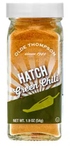 A clear spice jar labeled Olde Thompson since 1944 contains 1.9 oz (54g) of Hatch Green Chili powder. The label features an image of a green chili pepper and a sunburst design.