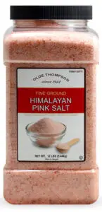 A large, clear plastic container labeled Olde Thompson since 1941 holds fine ground Himalayan pink salt. The label includes an image of a bowl of the salt and specifies the net weight as 12 lbs (5.44 kg).