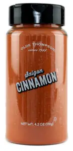 A clear plastic jar labeled Olde Thompson Saigon Cinnamon with a black lid. It contains 4.2 oz (119g) of ground cinnamon. The label notes since 1944.