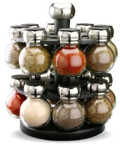 A rotating spice rack with labeled glass jars filled with various spices like oregano, chili powder, celery seed, paprika, garlic salt, black pepper, and thyme. The rack has a sleek, modern design with metallic lids.