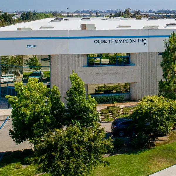 Exterior view of a building labeled Olde Thompson Inc. with trees and shrubs in front. The structure is white with large windows and has the number 2300 on it. A parking lot is partially visible in the background.