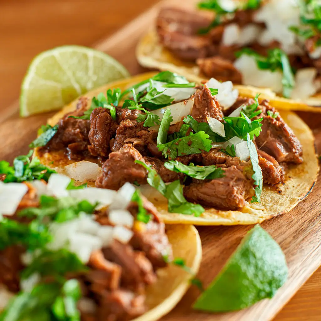 Close-up of three tacos filled with seasoned meat, topped with chopped onions and fresh cilantro, served on a wooden board. Lime wedges are placed beside the tacos for garnish.