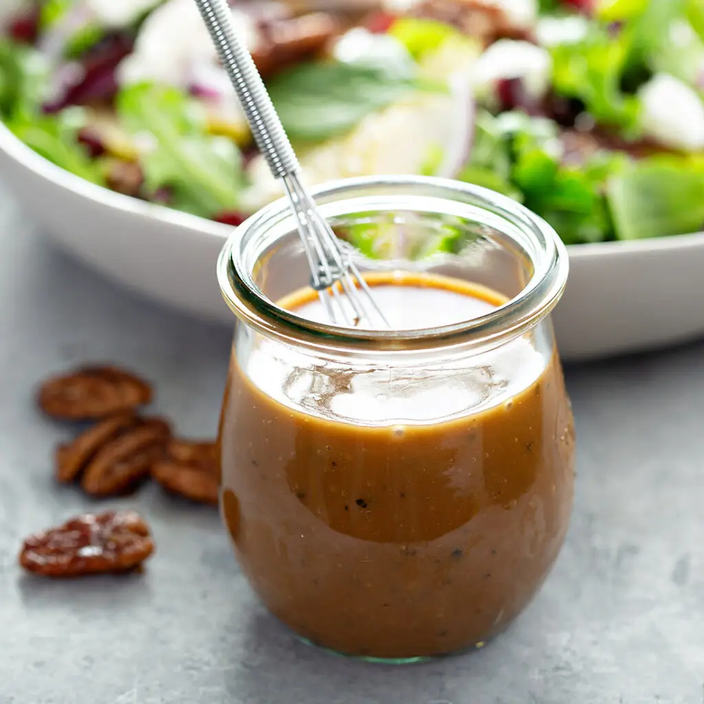 A glass jar filled with creamy brown salad dressing, with a silver spoon inside. The background features a blurred bowl of salad with leafy greens and visible pecans on the table.