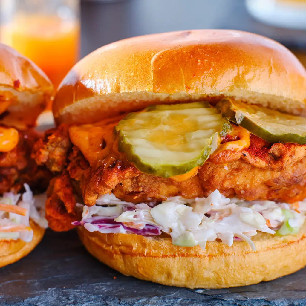 A close-up of a fried chicken sandwich with crispy golden chicken, creamy coleslaw, and pickle slices on a glossy brioche bun. The sandwich is served on a slate surface, with a blurred beverage in the background.