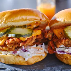 Two fried chicken sandwiches with coleslaw, pickles, and melted cheese on shiny buns. A blurred drink is in the background. The sandwiches are on a dark surface, highlighting their crispy texture and vibrant colors.