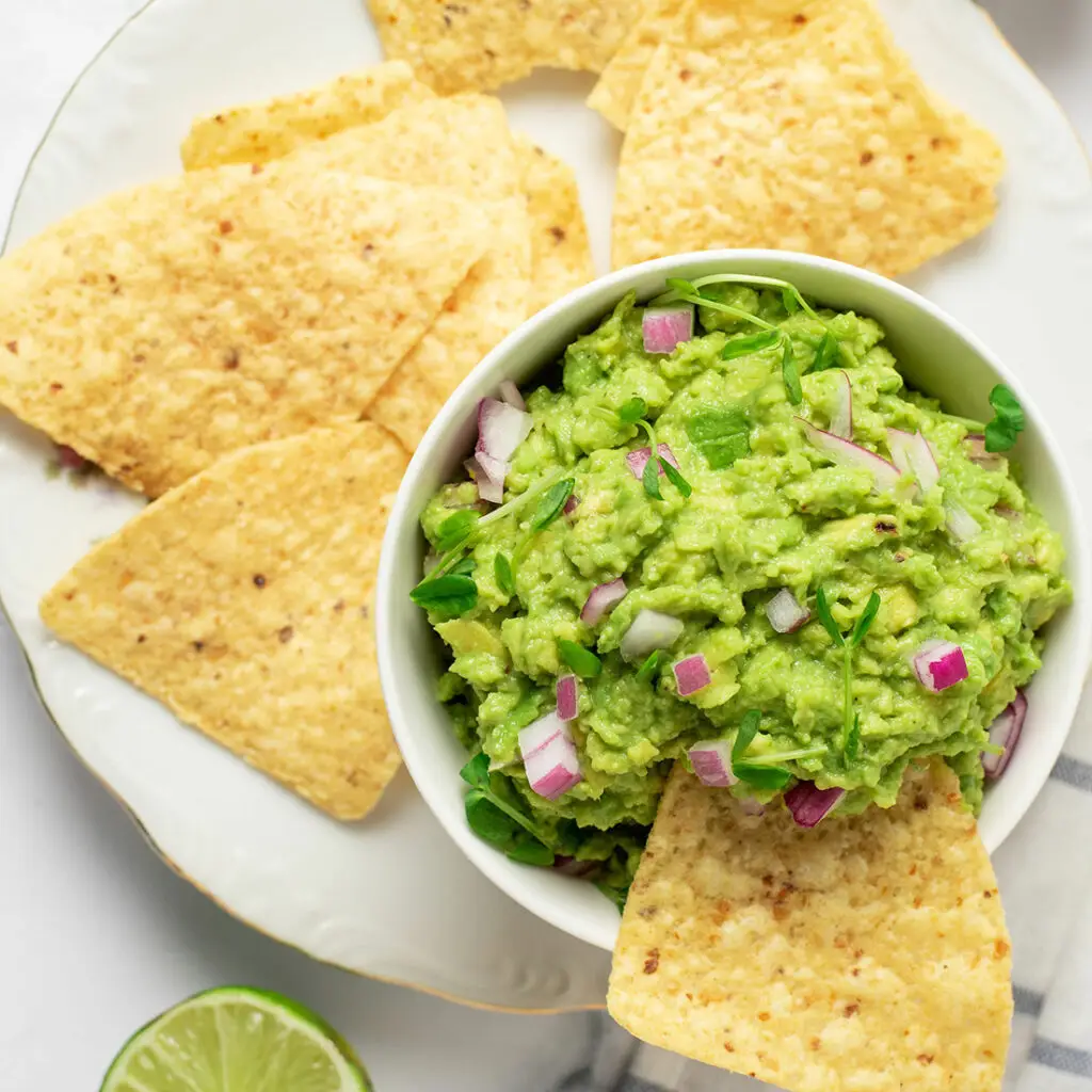 A bowl of guacamole topped with chopped red onions and cilantro, accompanied by tortilla chips on a white plate. A sliced lime is visible in the corner.