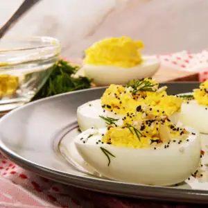 A plate of deviled eggs garnished with dill and spices sits on a table. In the background, a glass bowl with more filling and a deviled egg are visible. The table is covered with a red and white patterned cloth.