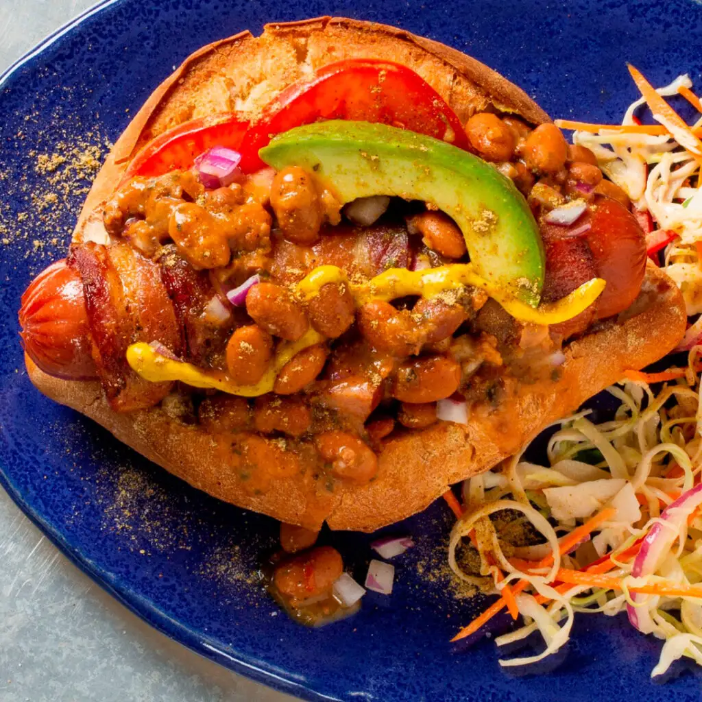 A hot dog in a bun topped with baked beans, avocado slice, diced onions, mustard, and tomato slices on a blue plate. Accompanied by a side of coleslaw.