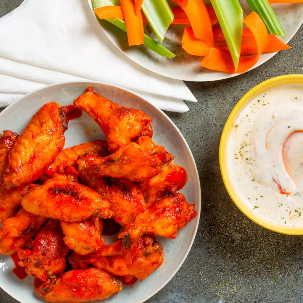 A plate of spicy buffalo chicken wings with a side of dipping sauce in a yellow bowl. Sliced carrots and celery are on a separate plate with a white napkin underneath. The background is a textured gray surface.