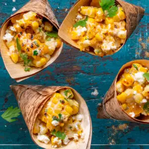 Four bamboo leaf cones filled with roasted corn kernels, crumbled cheese, and herbs are arranged on a rustic blue wooden surface. The dish is garnished with cilantro and a sprinkle of spices.