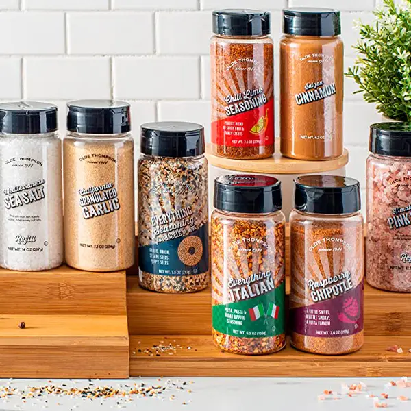 A variety of spice containers displayed on wooden platforms. The collection includes sea salt, garlic, everything seasoning, Italian seasoning, cinnamon, and more. The background has a white tiled wall and a small plant.