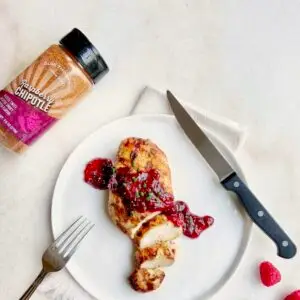 Grilled chicken breast topped with raspberry sauce on a white plate, accompanied by a knife and fork. A jar of raspberry chipotle seasoning is next to the plate. A single raspberry rests nearby on a light surface.