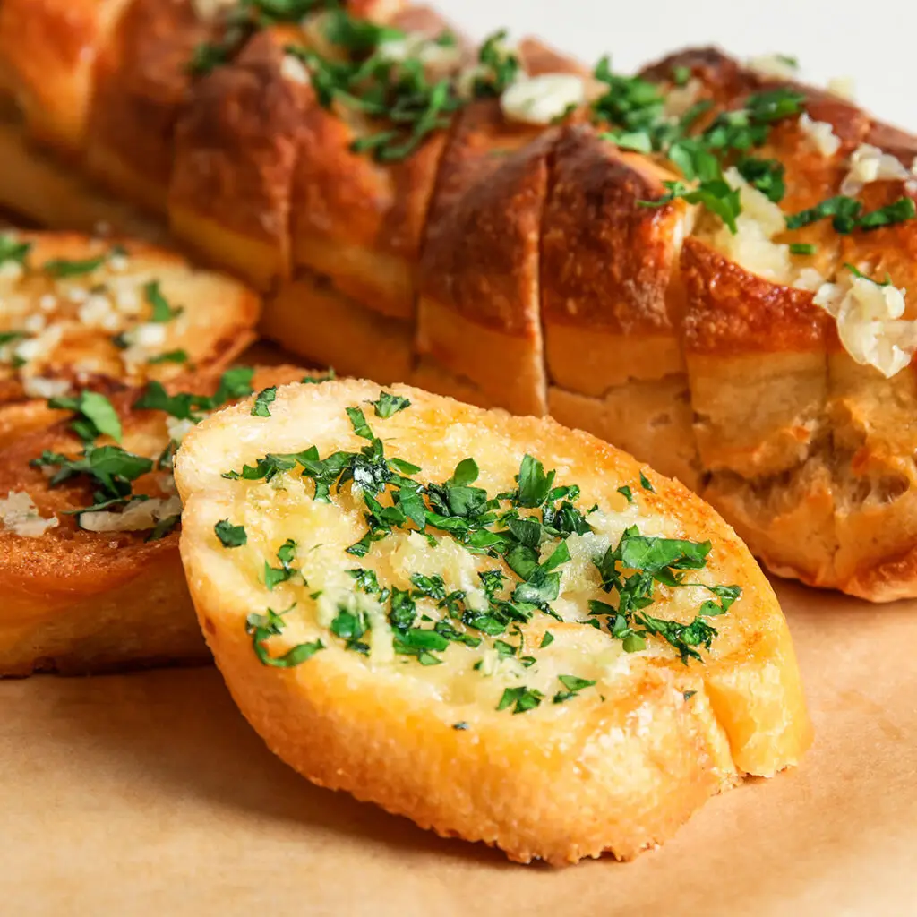 A loaf of garlic bread with a slice cut and placed in front. The bread is golden brown with a topping of melted butter, minced garlic, and chopped parsley. It rests on a wooden surface.