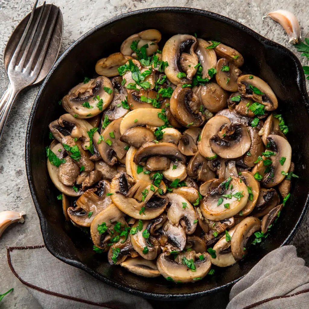 A skillet filled with sautéed mushrooms garnished with chopped parsley. Two forks and cloves of garlic are positioned beside the skillet. The background features a rustic, stone-like surface.