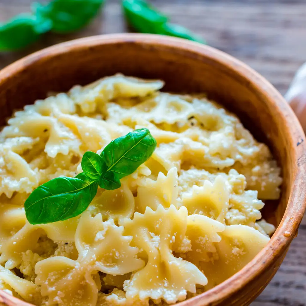 A wooden bowl filled with creamy Farfalle pasta, garnished with two fresh basil leaves. The pasta is sprinkled with grated cheese. The background includes basil leaves on a wooden surface.