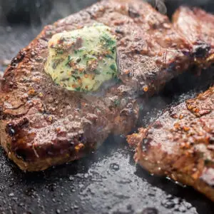 Close-up of two sizzling steaks on a grill, topped with a melting dollop of herb butter. The meat is seasoned with visible cracked black pepper and herbs, with steam rising, indicating they are cooking. The steaks are juicy and well-seared.