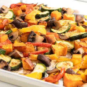 A close-up of roasted vegetables on a baking sheet, including zucchini, bell peppers, mushrooms, and sweet potato cubes. The vegetables are seasoned, lightly browned, and mixed together, creating a colorful and appetizing dish.