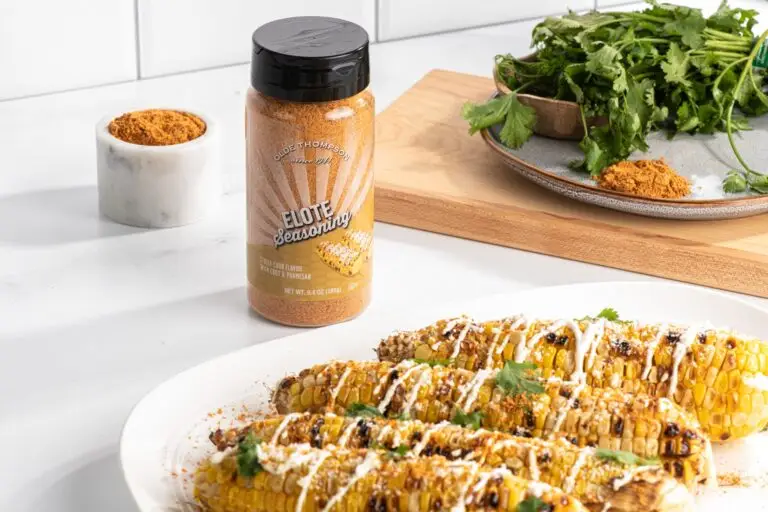 Grilled corn on a white plate garnished with seasoning and cilantro, drizzled with sauce. A jar of Eliot Seasoning stands on a counter near a small bowl of seasoning and a wooden board with fresh cilantro and more seasoning.