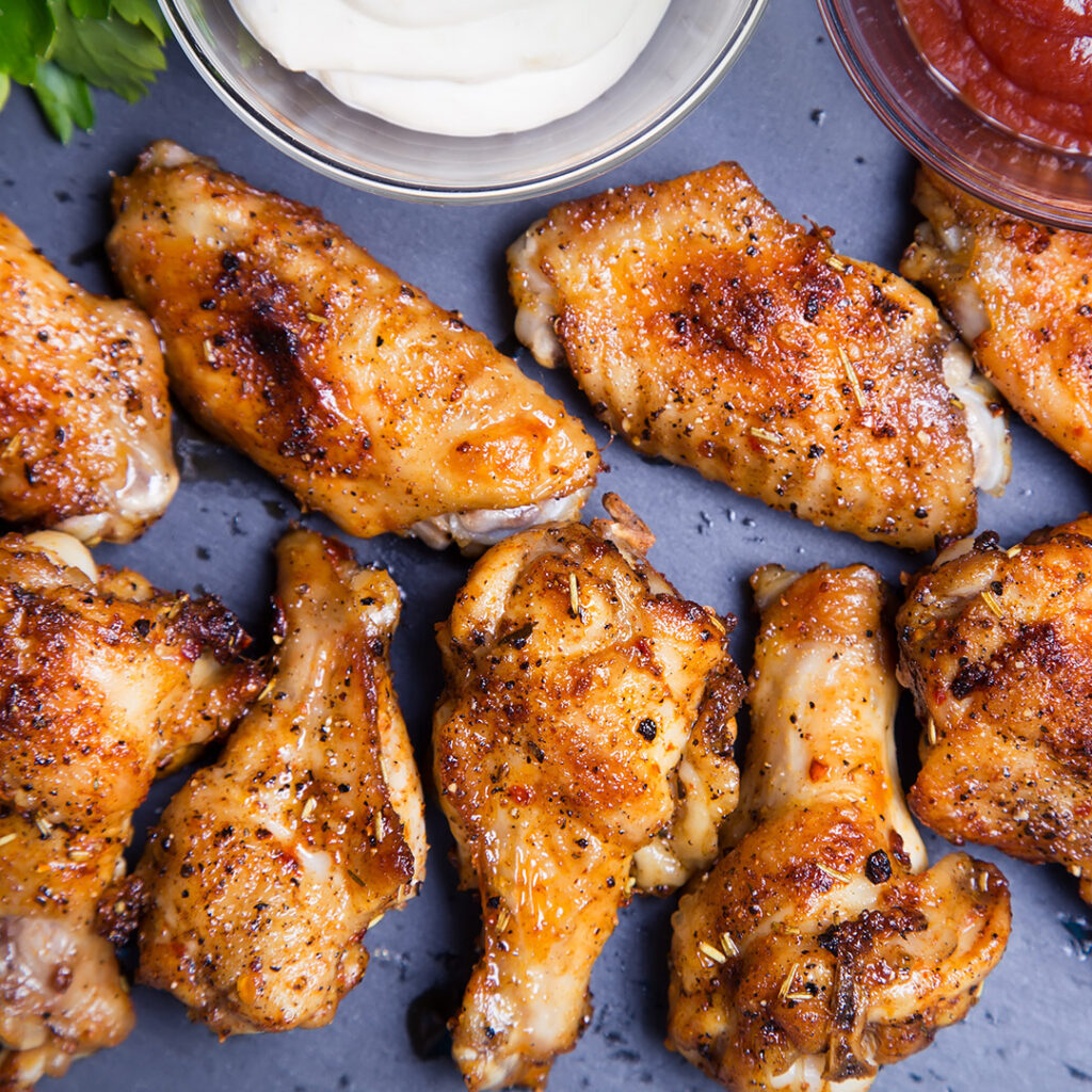 A close-up of crispy baked chicken wings dusted with Cacio e Pepe seasoning, arranged on a dark surface, with bowls of creamy dipping sauce and ketchup in the background.