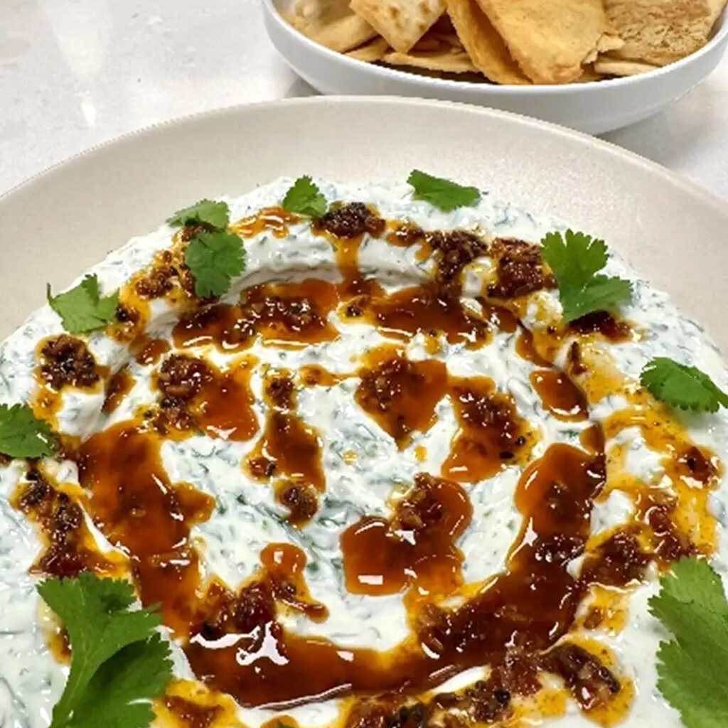 A bowl of creamy yogurt dip with herbs, drizzled with a reddish-brown spicy oil and garnished with fresh cilantro, served alongside pita chips and a side of Roasted and Tomato Garlic Salsa in the background.