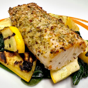 A seasoned, baked fish fillet with pesto seasoning sits atop grilled yellow squash and wilted spinach, garnished with herbs, on a white plate with an orange sauce accent.
