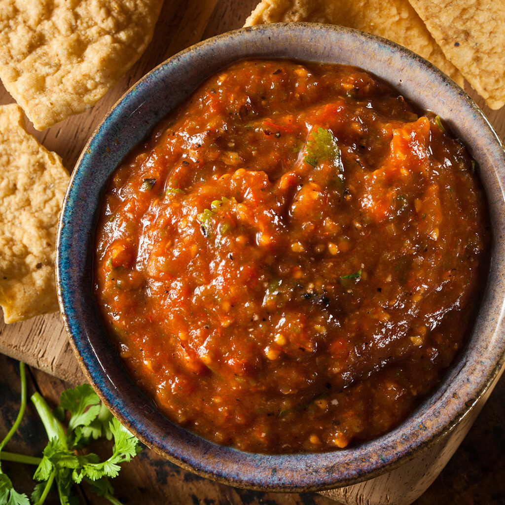 A bowl of chunky Roasted and Tomato Garlic Salsa sits on a wooden surface, surrounded by tortilla chips and fresh cilantro. The vibrant salsa features visible pieces of tomato, garlic, and green herbs.