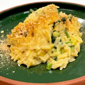 A serving of creamy orzo pasta mixed with green vegetables and a hint of Smoked Onion & Garlic Seasoning is topped with a golden breadcrumb crust and presented on a dark green plate.