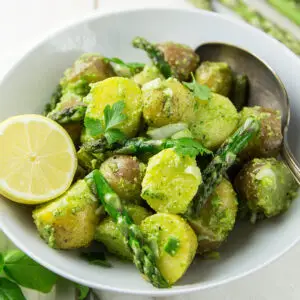 A white bowl filled with potato salad featuring halved baby potatoes, asparagus, fresh herbs, and a vibrant pesto seasoning green dressing. A lemon half and a serving spoon rest on the side.
