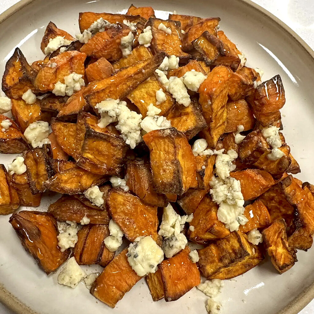 A plate of roasted sweet potato chunks, seasoned with Smoked Onion & Garlic Seasoning and topped with crumbled blue cheese, served on a round beige dish.