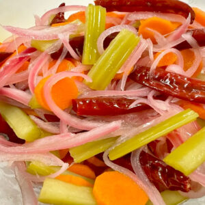 A close-up of Mexican pickled vegetables, featuring sliced pink onions, carrots, celery sticks, and whole dried red chilies, paired perfectly with Roasted and Tomato Garlic Salsa. The vegetables appear crisp and vibrant in color.
