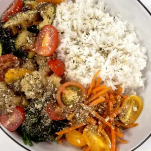 A plate with white rice, roasted mixed vegetables including broccoli, zucchini, cauliflower, and cherry tomatoes, and a side of shredded carrots and yellow tomatoes, all garnished with herbs and pesto seasoning.